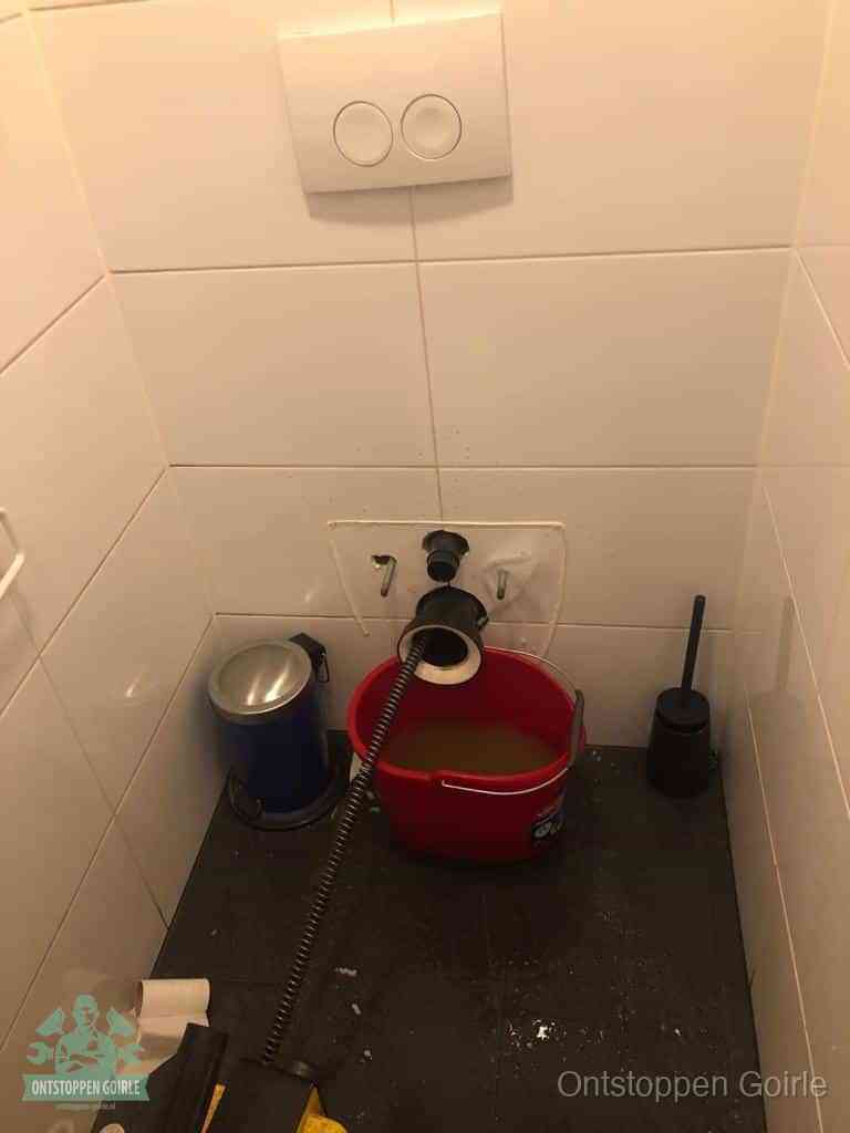 Een ontstopte wand-wc met verwijderde toiletpot waar een ontstoppingsveer in de wanduitgang verdwijnt, het achterhalen waar de verstopping zit in Goirle