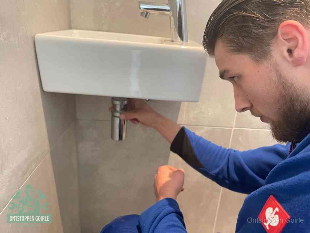 Een monteur draait met de hand de zwanenhalssifon onder een fonteinwasbak in de toiletruimte los om de bron van rioollucht in Goirle te vinden